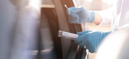 Zdjęcie osoby w stroju ochronnym stojącej obok otwartego okna w samochodzie która wyciąga ręce z próbówką aby pobrać wymaz od osoby siedzącej w samochodzie