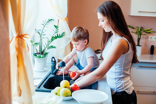 Zdjęcie kobiety stojącej przy zlewie w kuchni, myjącej warzywa. Obok niej, na krawędzi zlewu siedzi dziecko i pomaga myć warzywa