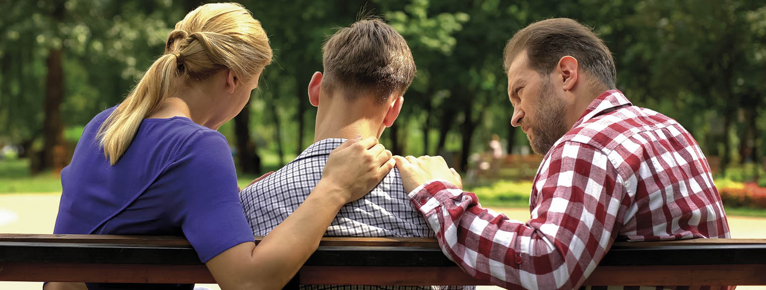 W parku na ławce siedzą 3 osoby. 2 osoby na brzegach ławki(matka i ojciec) trzymają ręce na barkach chłopaka po środku. Zdjęcie jest zrobione z tyłu ławki.